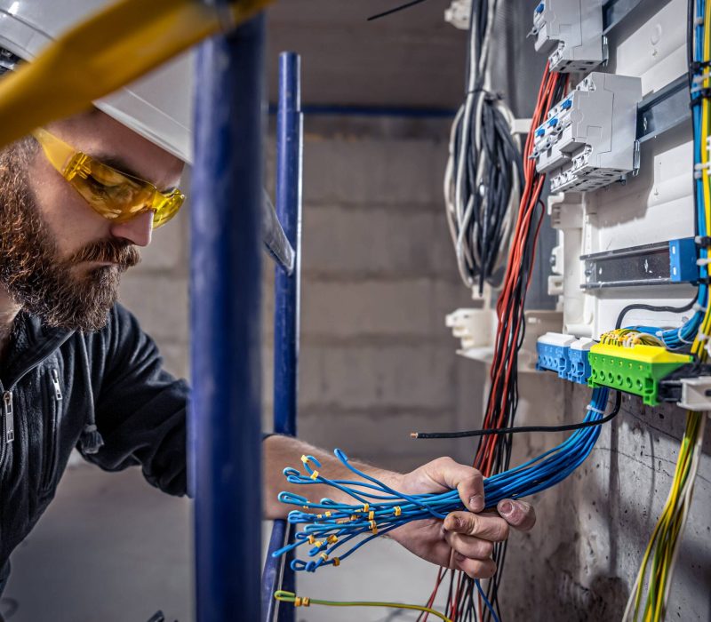 A male electrician works in a switchboard with an electrical connecting cable, connects the equipment with tools.